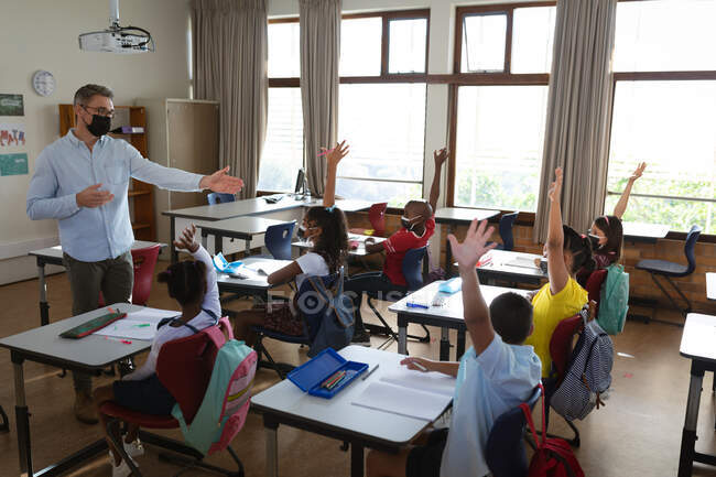 Grupo de diversos estudiantes con máscaras faciales levantando las manos en la clase en la escuela. higiene y distanciamiento social en la escuela durante la pandemia de covid 19 - foto de stock