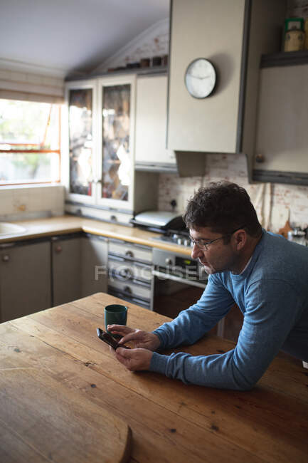Homem caucasiano na cozinha de pé à mesa e usando smartphone. passar tempo livre em casa. — Fotografia de Stock