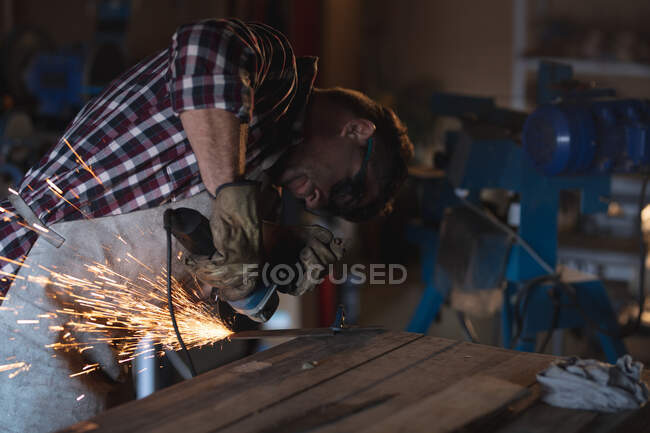 Caucasian male knife maker wearing apron and glasses, using angle grinder in workshop. independent small business craftsman at work. — Stock Photo