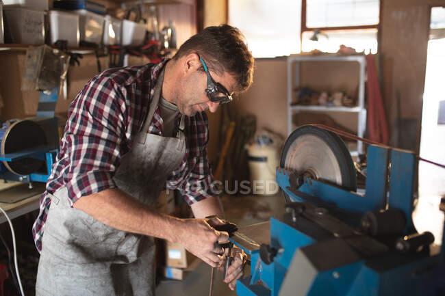 Caucasian male knife maker wearing apron and glasses , making knife in workshop. independent small business craftsman at work. — Stock Photo