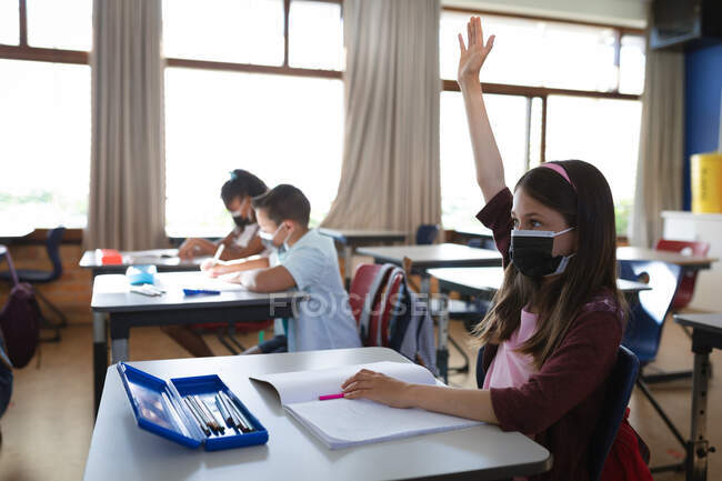 Kaukasisches Mädchen mit Mundschutz hebt die Hand, während sie in der Schulklasse auf ihrem Schreibtisch sitzt. Hygiene und soziale Distanzierung in der Schule während der covid 19 Pandemie — Stockfoto