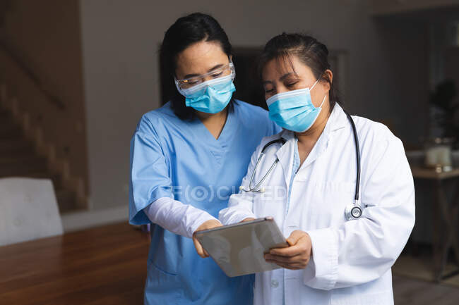 Dos mujeres médicas asiáticas usando mascarillas en casa usando tableta. atención médica y fisioterapia médica tratamiento. - foto de stock