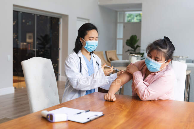Medico asiatico di sesso femminile che indossa maschera facciale e vaccinare paziente femminile a casa. cure sanitarie e fisioterapiche. — Foto stock