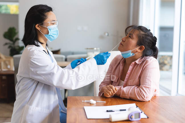 Médica asiática usando máscara facial e fazendo teste de esfregaço de paciente do sexo feminino em casa. tratamento de saúde e fisioterapia médica. — Fotografia de Stock