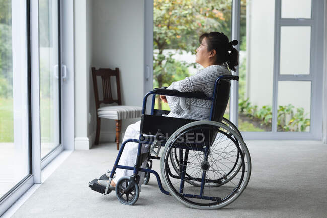 Asiatin sitzt im Rollstuhl und schaut zu Hause durchs Fenster. Gesundheitswesen und medizinische physiotherapeutische Behandlung. — Stockfoto