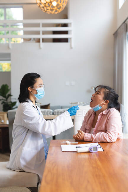 Азиатка-врач в маске для лица и сдаёт мазок пациентке дома. медицинское и физиотерапевтическое лечение. — стоковое фото