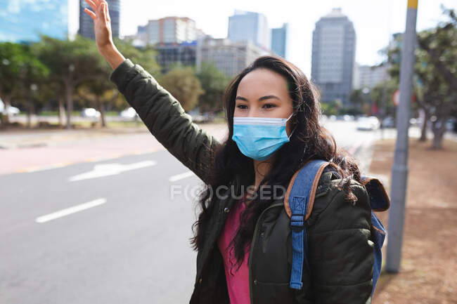 Mujer asiática usando mascarilla de pie junto al camino llamando a un taxi. mujer joven independiente fuera y alrededor de la ciudad durante coronavirus covid 19 pandemia. - foto de stock