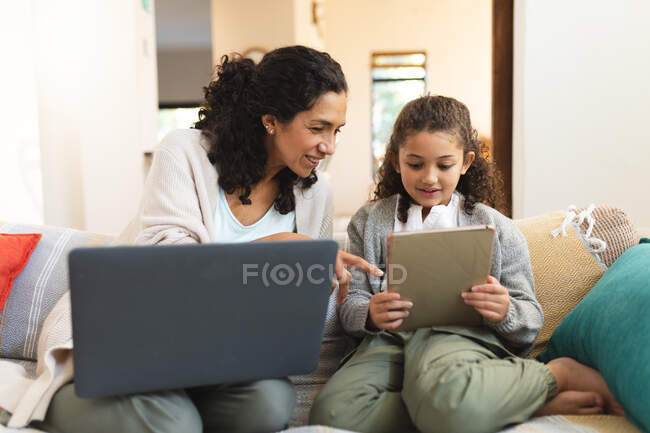 Sonriente madre de raza mixta e hija sentadas en el sofá, utilizando el portátil y la tableta. estilo de vida doméstico y pasar tiempo de calidad en casa. - foto de stock