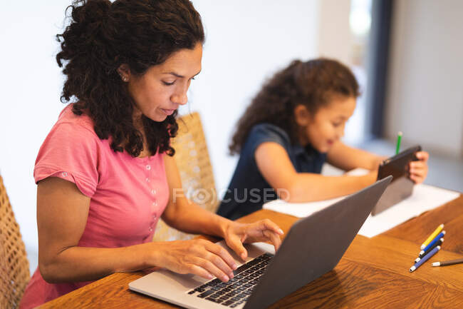 Mixed race mother and daughter sitting at table, using laptop and tablet. domestic lifestyle and spending quality time at home. — Stock Photo