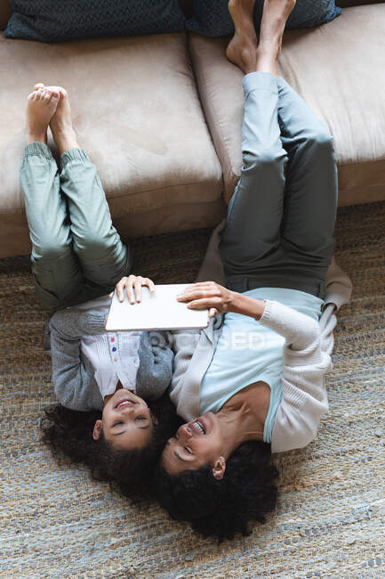 Mixed race mother and daughter lying on carpet, using tablet in living room. domestic lifestyle and spending quality time at home. — Stock Photo