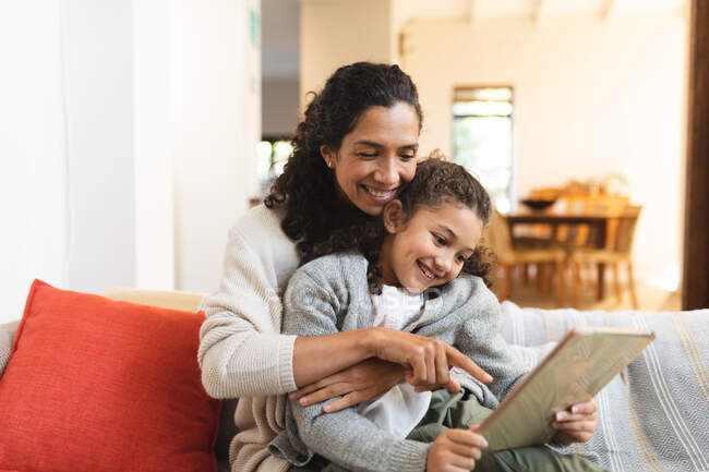 Sorrindo mestiço mãe e filha sentados no sofá, usando tablet. estilo de vida doméstico e passar tempo de qualidade em casa. — Fotografia de Stock