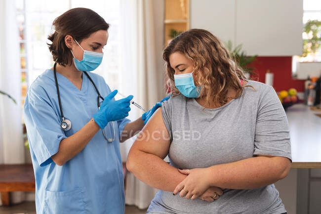 Kaukasische Ärztin mit Gesichtsmaske impft Patientin zu Hause. Hausbesuche während der Coronavirus-Pandemie 19. — Stockfoto