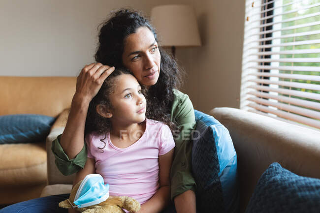 Mixed race mother and daughter sitting on sofa, embracing and looking through window. domestic lifestyle and spending quality time at home. — Stock Photo
