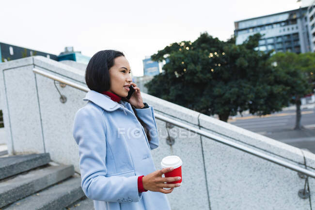 Mujer asiática sosteniendo el café para llevar y utilizando el teléfono inteligente en las escaleras. mujer joven independiente fuera y alrededor de la ciudad. - foto de stock