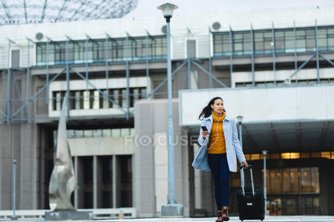 Mujer asiática sosteniendo teléfono inteligente y caminando con la maleta en la calle. mujer joven independiente fuera y alrededor de la ciudad. — Stock Photo