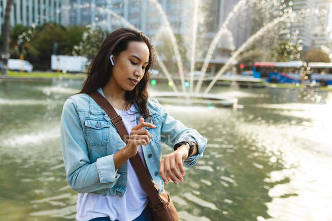 Азиатка сидит и проверяет умные часы в солнечном парке. независимая молодая женщина в городе. — стоковое фото