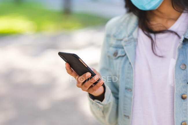 Frau mit Mundschutz mit Smartphone im sonnigen Park. Unabhängige junge Frau während der Coronavirus-Pandemie 19 in der Stadt unterwegs. — Stockfoto