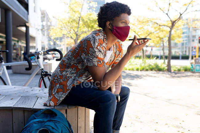 Uomo afroamericano in città che indossa faccia seduta e utilizzando smartphone. nomade digitale in movimento, in giro per la città durante il coronavirus covid 19 pandemia. — Foto stock