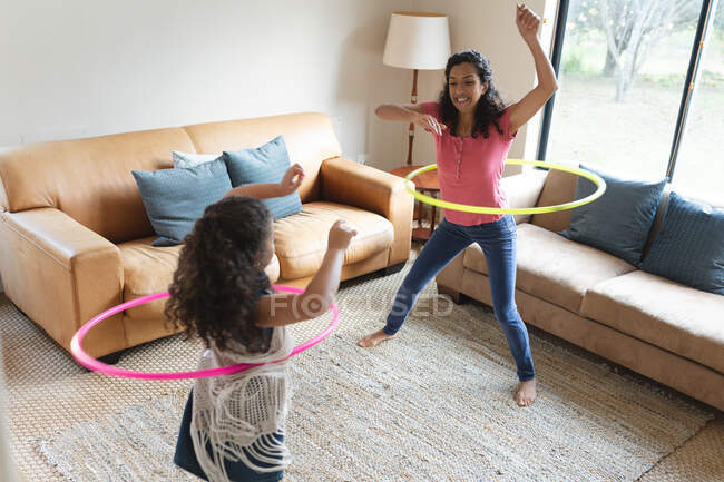 Feliz madre de raza mixta e hija jugando con hula hoop en la sala de estar. estilo de vida doméstico y pasar tiempo de calidad en casa. - foto de stock