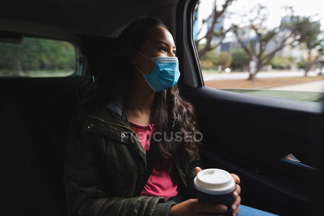 Азиатка в маске сидит в такси, держит кофе на вынос. Независимая молодая женщина вне и около в городе во время коронавируса ковид 19 пандемии. — стоковое фото