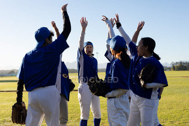 Разнообразная группа счастливых женщин-бейсболистов поднимает руки на солнечном бейсбольном поле перед игрой. женская бейсбольная команда, спортивные тренировки, сплоченность и приверженность. — стоковое фото