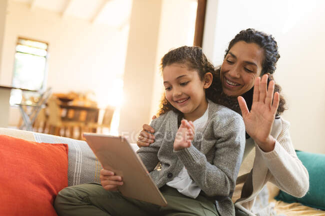 Smiling mixed race mother and daughter sitting on sofa using tablet, having video call. domestic lifestyle and spending quality time at home. — Stock Photo