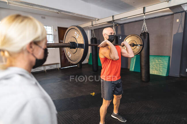 Kaukasische Trainerin, die Mann beim Training im Fitnessstudio anweist, Gesichtsmasken zu tragen und Gewichte zu heben. Kraft- und Fitnesstraining für das Boxen während der Coronavirus-Pandemie 19. — Stockfoto
