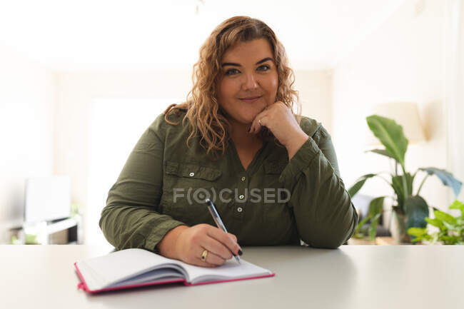 Retrato de mulher caucasiana fazendo uma chamada de vídeo, tomando notas e sorrindo. estilo de vida doméstico, passar o tempo livre em casa. — Fotografia de Stock