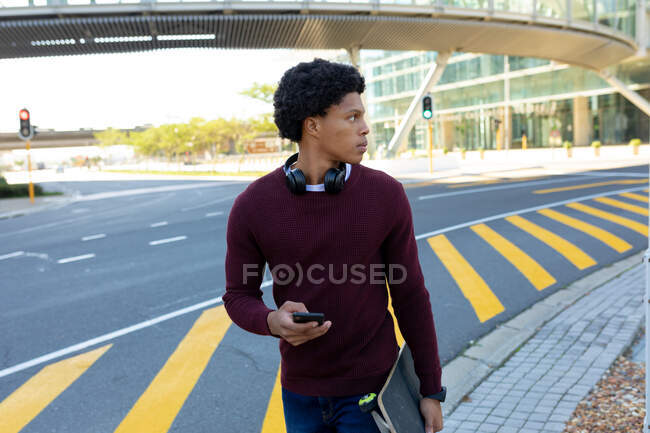 Homme afro-américain en ville utilisant un smartphone et tenant skateboard. nomade numérique en déplacement, en déplacement dans la ville. — Photo de stock