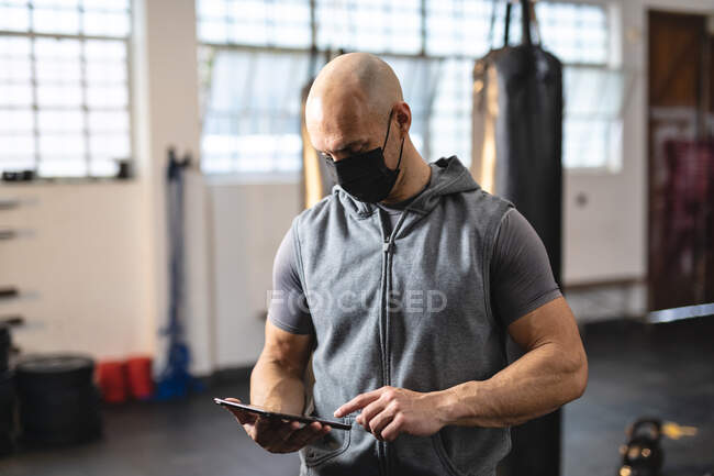 Caucasian male trainer wearing face mask, using tablet. strength and fitness cross training for boxing during coronavirus covid 19 pandemic. — Stock Photo
