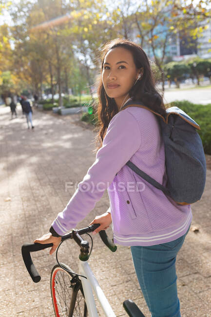 Portrait of smiling asian woman wheeling bike and looking at camera in sunny park. independent young woman out and about in the city. — Stock Photo