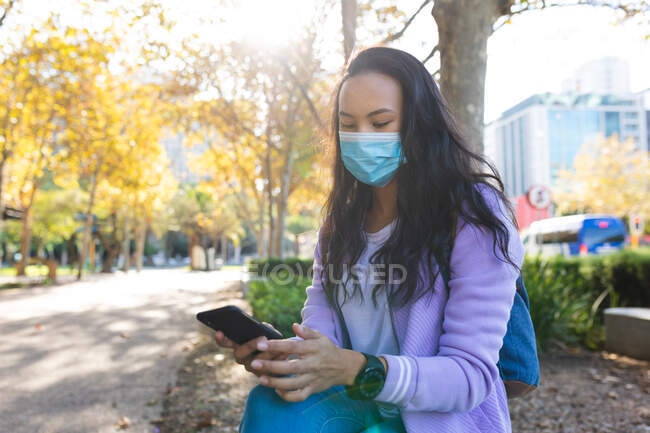 Азійка, одягнена в маску обличчя, користується смартфоном у сонячному парку. Незалежна молода жінка, яка живе в місті під час коронавірусу (19 пандемії). — стокове фото