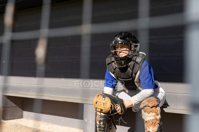 Jugadora de béisbol caucásica sentada en el banco con casco de receptor y ropa protectora. equipo femenino de béisbol, preparado y esperando el juego. - foto de stock