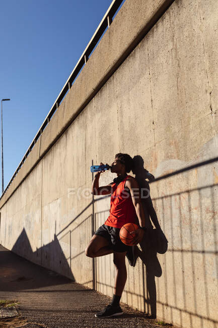 Fit african american man exercising in city holding basketball and drinking water in the street. fitness and active urban outdoor lifestyle. — Stock Photo