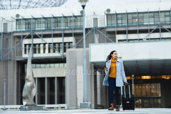 Mujer asiática sosteniendo teléfono inteligente y caminando con la maleta en la calle. mujer joven independiente fuera y alrededor de la ciudad. - foto de stock