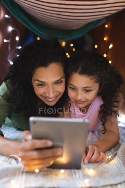 Retrato de madre e hija feliz de raza mixta usando un portátil en una tienda improvisada. estilo de vida doméstico y pasar tiempo de calidad en casa. - foto de stock