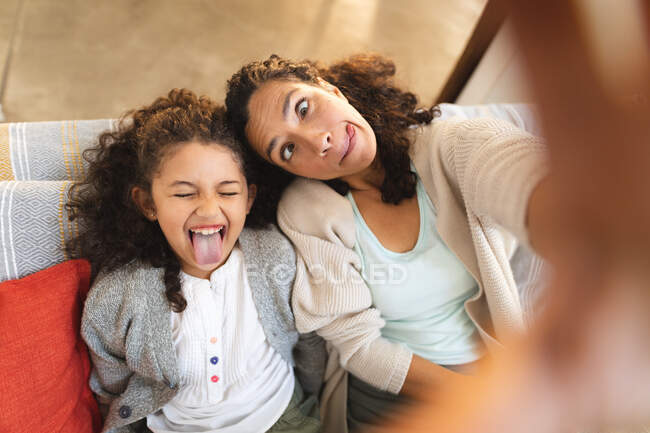 Raza mixta madre e hija sentadas en el sofá haciendo caras graciosas, tomando selfie. estilo de vida doméstico y pasar tiempo de calidad en casa. - foto de stock