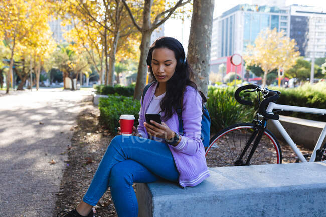 Mujer asiática con audífonos usando smartphone y sosteniendo café para llevar en el soleado parque. mujer joven independiente fuera y alrededor de la ciudad. - foto de stock