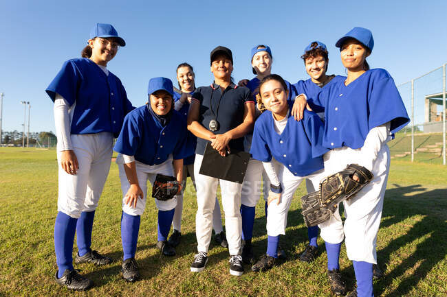 Портрет разнообразной группы улыбающихся женщин-бейсболистов и тренера, стоящих на солнечном поле. женская бейсбольная команда, спортивные тренировки, сплоченность и приверженность. — стоковое фото