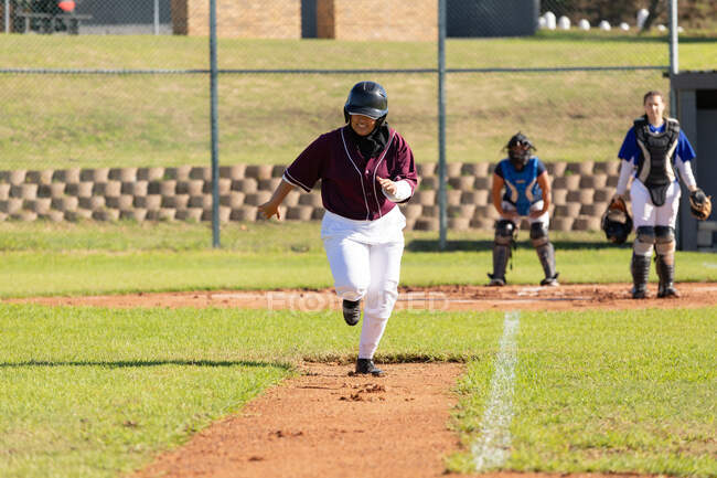 Jogadora de beisebol mista em campo de beisebol ensolarado correndo entre bases durante o jogo. time de beisebol feminino, treinamento esportivo e táticas de jogo. — Fotografia de Stock