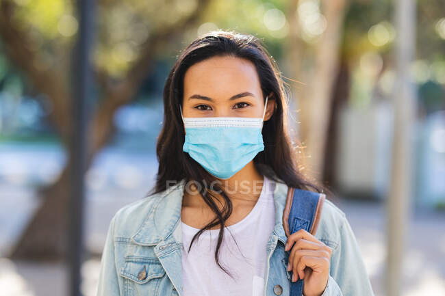 Ritratto di donna asiatica che indossa maschera viso guardando la fotocamera nel parco soleggiato. indipendente giovane donna in giro per la città durante coronavirus covid 19 pandemia. — Foto stock