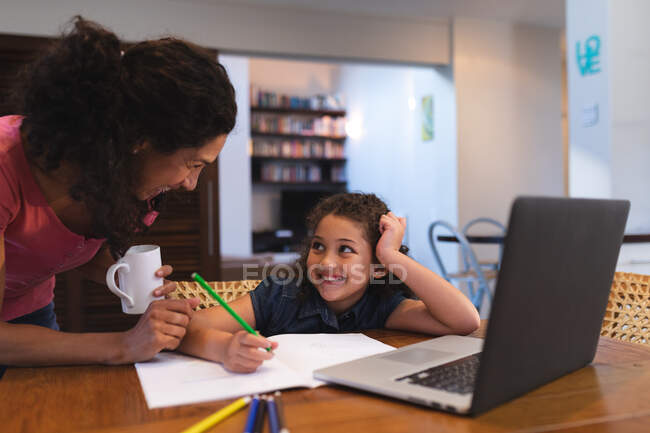 Sonriente madre de raza mixta sosteniendo café, ayudando a su hija a hacer la tarea. estilo de vida doméstico y pasar tiempo de calidad en casa. - foto de stock