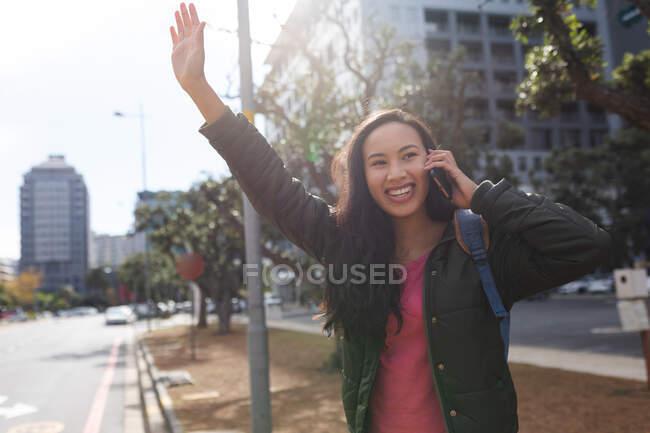 Азійська жінка, яка стоїть на дорозі, вітає таксі, розмовляючи по смартфону. Незалежна молода жінка у місті.. — стокове фото