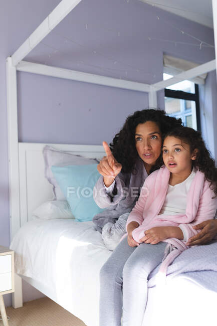 Glückliche gemischte Rasse Mutter und Tochter sitzen auf dem Bett, umarmen und schauen durch das Fenster. Lebensstil und hochwertige Zeit zu Hause verbringen. — Stockfoto