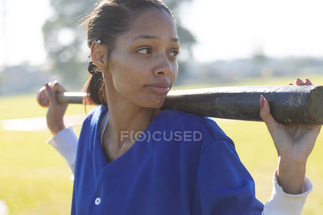 Jogadora de beisebol mista segurando taco de beisebol nos ombros olhando para longe no campo de beisebol. time de beisebol feminino, preparado e esperando o jogo. — Fotografia de Stock