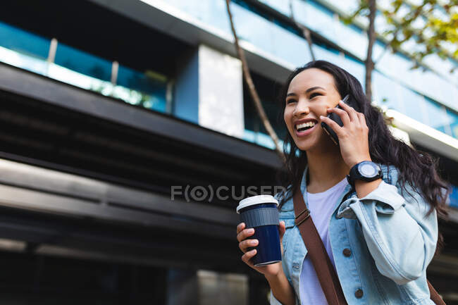Mujer asiática usando smartphone y sosteniendo café para llevar en la calle. mujer joven independiente fuera y alrededor de la ciudad. - foto de stock