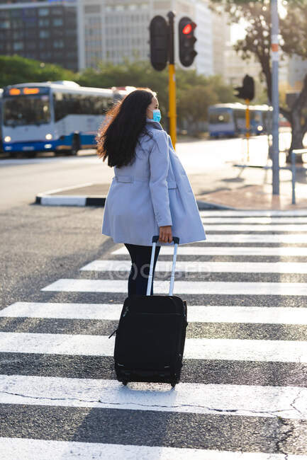 Азиатка в маске и переходит дорогу с чемоданом. Независимая молодая женщина вне и около в городе во время коронавируса ковид 19 пандемии. — стоковое фото