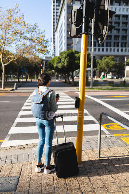 Азиатка переходит дорогу с чемоданом и держит кофе на вынос. независимая молодая женщина в городе. — стоковое фото