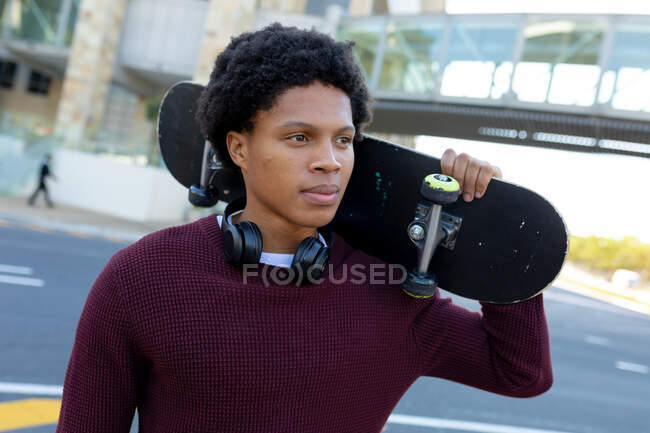 Hombre afroamericano en la ciudad sosteniendo monopatín. nómada digital sobre la marcha, fuera y alrededor de la ciudad. - foto de stock