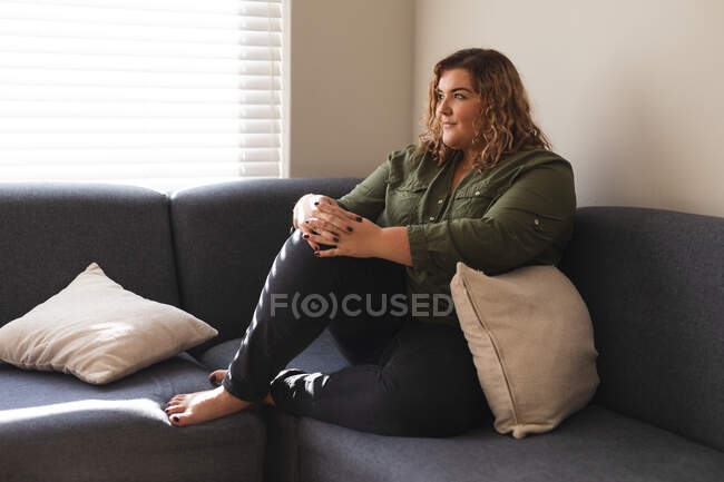 Mujer caucásica mirando hacia otro lado y sentada en el sofá. estilo de vida doméstico, pasar tiempo libre en casa. - foto de stock